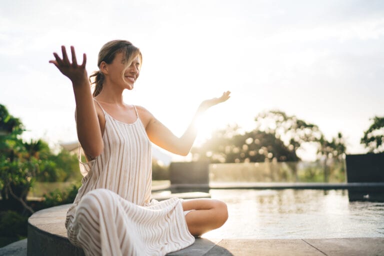 Young happy woman practicing yoga and meditating on swimming pool edge in yard of resort hotel. Tourism, vacation and weekend. Pretty caucasian girl. Sunset. Idyllic and calm lifestyle