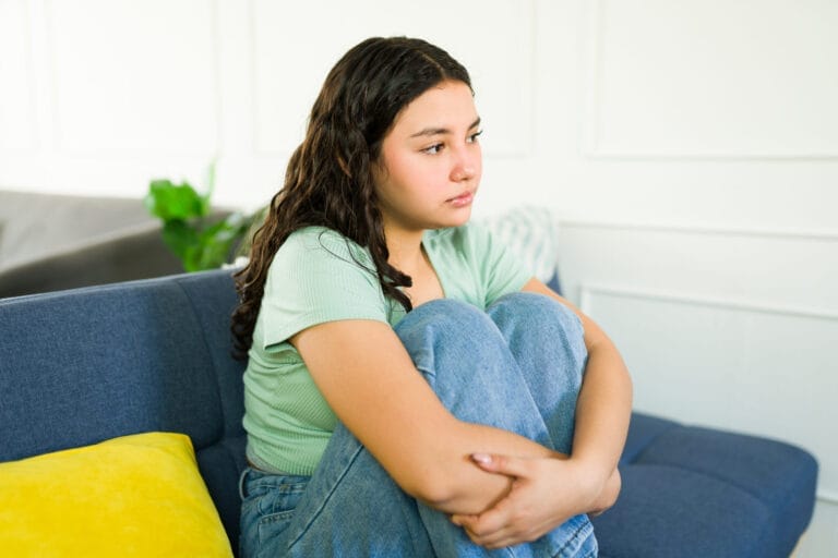 Sad teen girl sitting on sofa hugging knees and feeling lonely and depressed at home